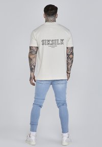 Kremasta majica kratkih rukava s crnim "SIKSILK" logotipom na leđima, u kombinaciji s svijetloplavim uskim trapericama i bijelim tenisicama.