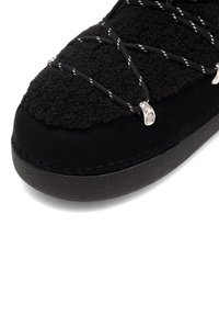 Fekete lábbeli, amelynek texturált felsőrésze bolyhos anyagból készült, sima fekete anyaggal keretezve; elasztikus fűzés fémes kiegészítőkkel.