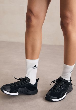 Person iført hvide Adidas mellemlange sokker og sorte sneakers med chunky såler, stående på en glat, lysefarvet gulv.