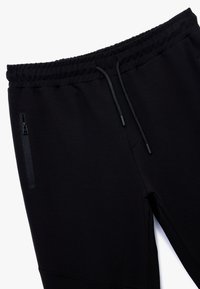 Sorte joggebukser fremstillet af et tekstureret stof, elastisk talje med snor og en sidelomme med lynlås til sikker opbevaring.
