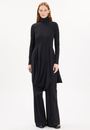 Donna che indossa una tunica nera asimmetrica a maniche lunghe sopra pantaloni neri a gamba larga, in piedi davanti a uno sfondo bianco uniforme.