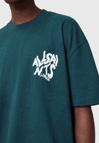 Tamsiai žalias medvilninis marškinėliai su baltu logo „ALLSAINTS LONDON“ kairėje krūtinės pusėje, laisvo kirpimo ir trumpomis rankovėmis.