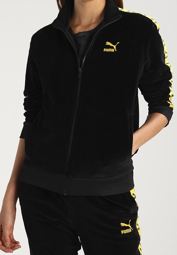 Personne portant un survêtement Puma noir avec un logo jaune et des bandes sur les manches et le pantalon, mains dans les poches de la veste.