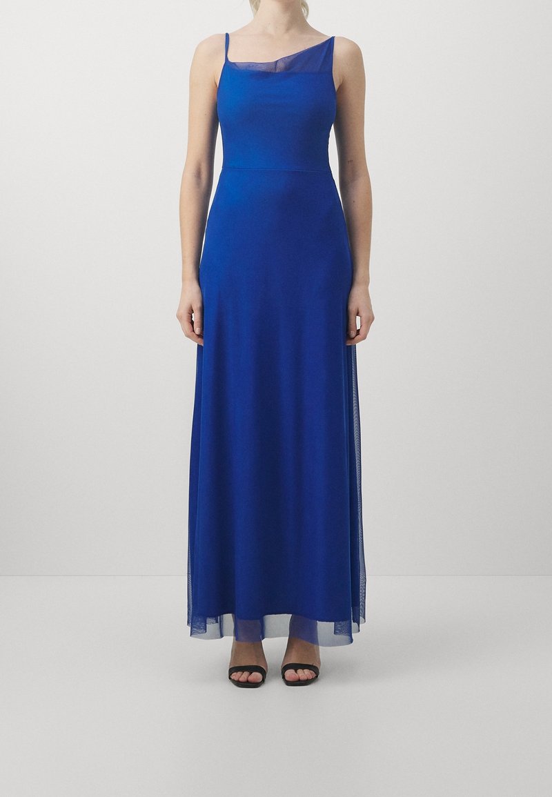 Femme portant une longue robe sans manches bleu roi avec un détail en tissu transparent au niveau du décolleté et de l'ourlet, associée à des talons ouverts noirs.