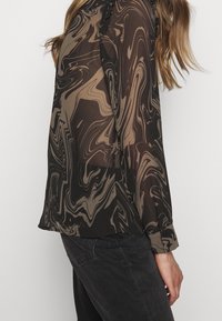 Vrouw in een lange mouw, zwart en bruin gemarmerd patroon doorzichtig blouse, gecombineerd met zwarte broek, zijaanzicht.