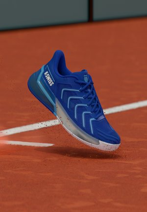 Chaussure de sport bleue K-Swiss soulevant de la poussière d'argile sur un court de tennis près d'une ligne de limite blanche.