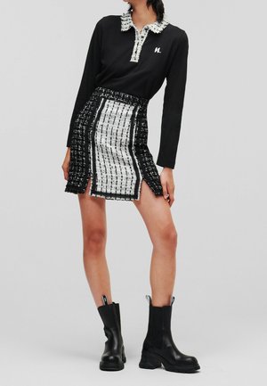 Nainen, joka pukeutunut mustaan pitkähihainen yläosa, jossa on teksturoidut valkoiset kaulukset, ja mustavalkoiseen tweed-minihameeseen, jossa on sivuhalkiot, yhdistettynä mustiin nilkkureihin.