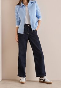 Mujer vestida con chaqueta de cremallera azul claro, camisa blanca, jeans de pierna ancha azul oscuro y zapatillas blancas, de pie contra una pared beige.