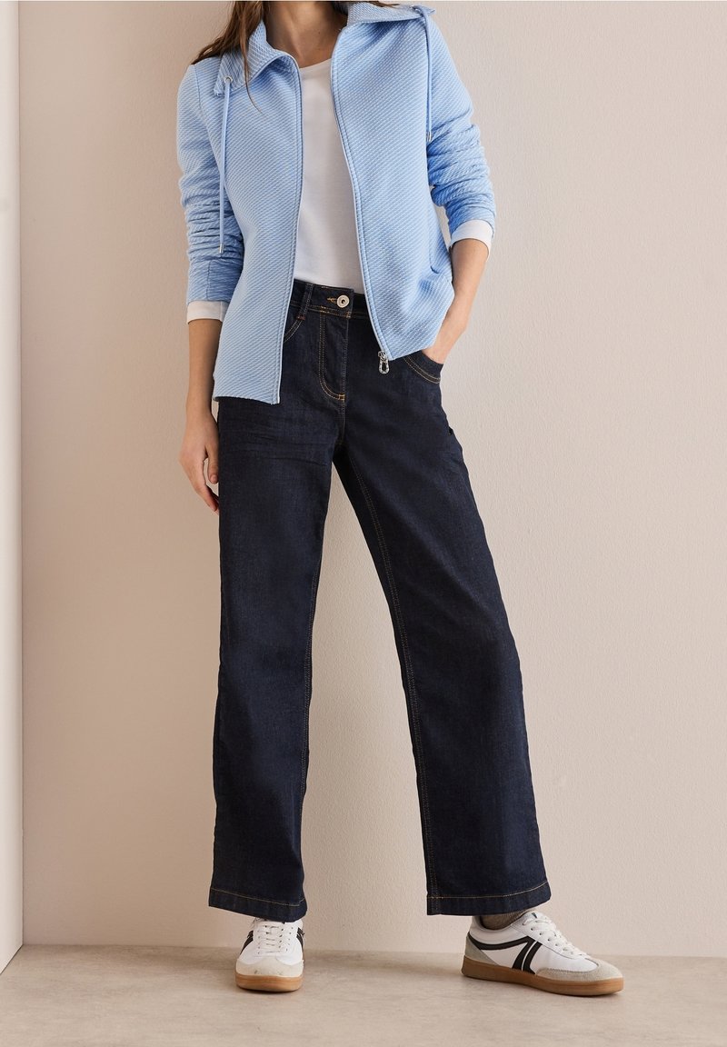 Mujer vestida con chaqueta de cremallera azul claro, camisa blanca, jeans de pierna ancha azul oscuro y zapatillas blancas, de pie contra una pared beige.