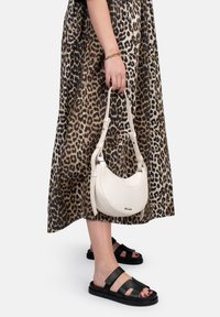 Sac à main en cuir blanc en forme de croissant avec une poignée ajustable, tenu par une personne portant une robe à imprimé léopard et des sandales noires.