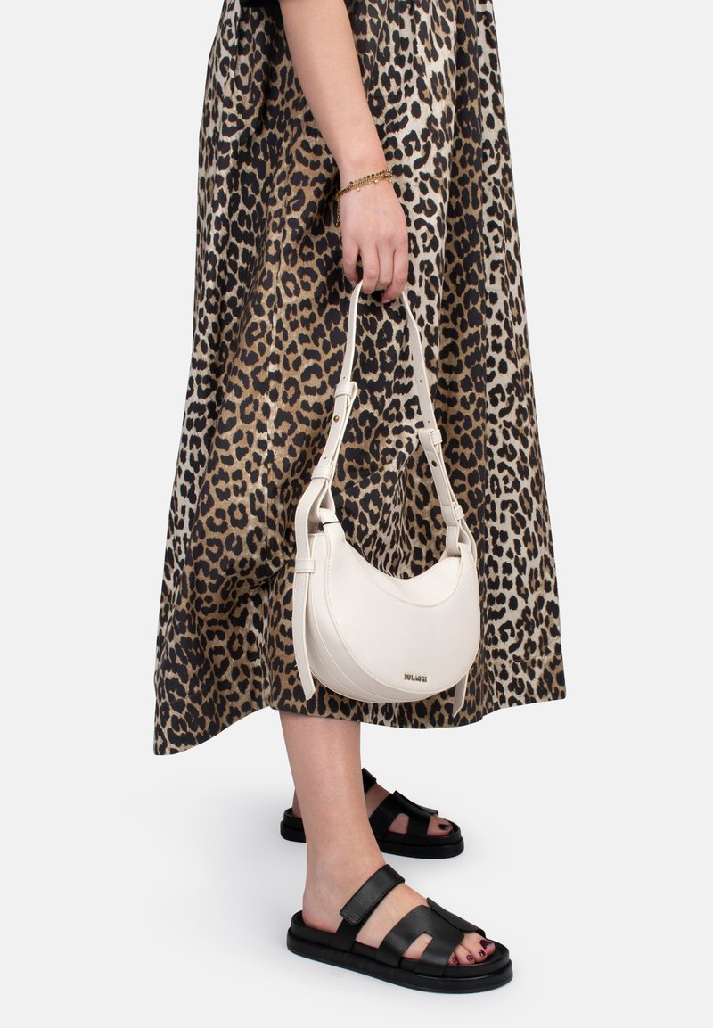 Sac à main en cuir blanc en forme de croissant avec une poignée ajustable, tenu par une personne portant une robe à imprimé léopard et des sandales noires.