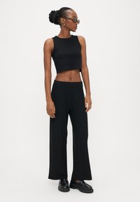 Črna kratka majica brez rokavov z gladko teksturo, združena z visokimi, širokimi črnimi hlačami z rebrastim vzorcem. Črni čevlji.