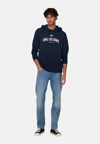Sudadera marinera con bolsillo canguro en la parte frontal, diseño de texto en blanco y rojo, combinada con jeans de un azul claro y zapatillas negras con suelas color crema.