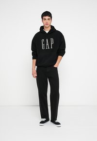 Felpa con cappuccio nera con scritta "GAP" bianca, taglio ampio, abbinata a pantaloni neri e sneakers nere. Outfit casual moderno dal design minimalista.