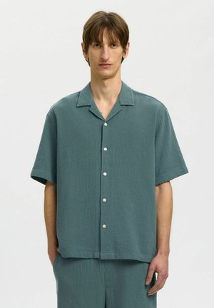 Jeune homme portant une chemise à manches courtes ample de couleur sarcelle avec boutons et un pantalon assorti, debout devant un fond blanc uni.