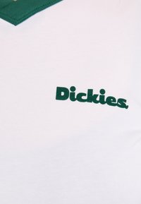 Balta marškinėliai su žaliu apykakle ir žaliu "Dickies" logotipu, užrašytu drąsiu, bešraifu šriftu ant krūtinės. Lygus medvilnės audinys.