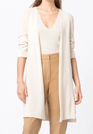 Femme portant un long cardigan beige sur un haut léger à col en V et un pantalon taille haute couleur sable sur fond blanc.