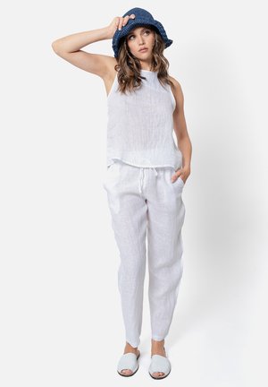 Witte linnen tanktop en broek met trekkoord; het model draagt ook een blauwe geweven bucket hat en lichte instapsandalen.