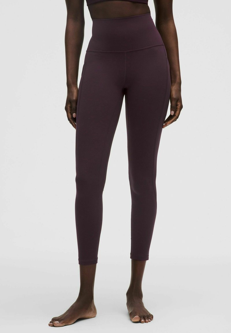 Frau, die hochtaillierte, dunkelviolette, verkürzte Leggings trägt, steht barfuß vor einem schlichten hellen Hintergrund.