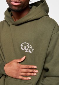 Felpa verde oliva in morbido misto cotone, con logo ricamato in argento sul petto. Include un ampio cappuccio e tasca frontale.