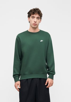 Fiatal férfi göndör hajjal, sötétzöld Nike pulóvert és fekete nadrágot visel, sima világos háttér előtt áll.