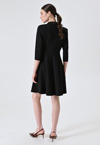Zwarte jurk tot de knie met driekwartmouwen, getailleerde bustier, uitlopende rok en een gladde textuur. Gecombineerd met bruine slingback hakken.