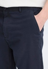 Chinos bleu marine avec une texture lisse, dotés de poches latérales, de passants de ceinture et d'une fermeture à bouton. Présentés avec un pull côtelé de couleur claire.