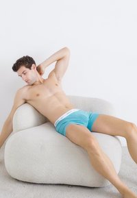 Turquoise boxer shorts with a white waistband worn by a person reclining on a textured, cream-colored, rounded sofa.