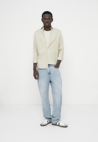 Ανοιχτοπράσινο πουκάμισο με κουμπιά, απαλό ροζ μπλουζάκι από μέσα, μπλε τζιν και μαύρα/λευκά αθλητικά παπούτσια· ουδέτερο φόντο, όρθια στάση.