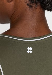 Ελαιοπράσινη αθλητική μπλούζα με λευκές λεπτομέρειες γύρω από τον λαιμό και ένα μικρό λευκό λογότυπο στο πίσω μέρος, που αναδεικνύει μια υφή υφάσματος.