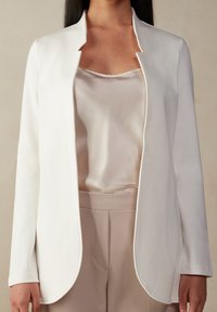 Bílý, lehký blejzr s jemnou texturou, otevřený vpředu a zaobleným lemem, nošený přes lehký saténový top a upravené béžové kalhoty.