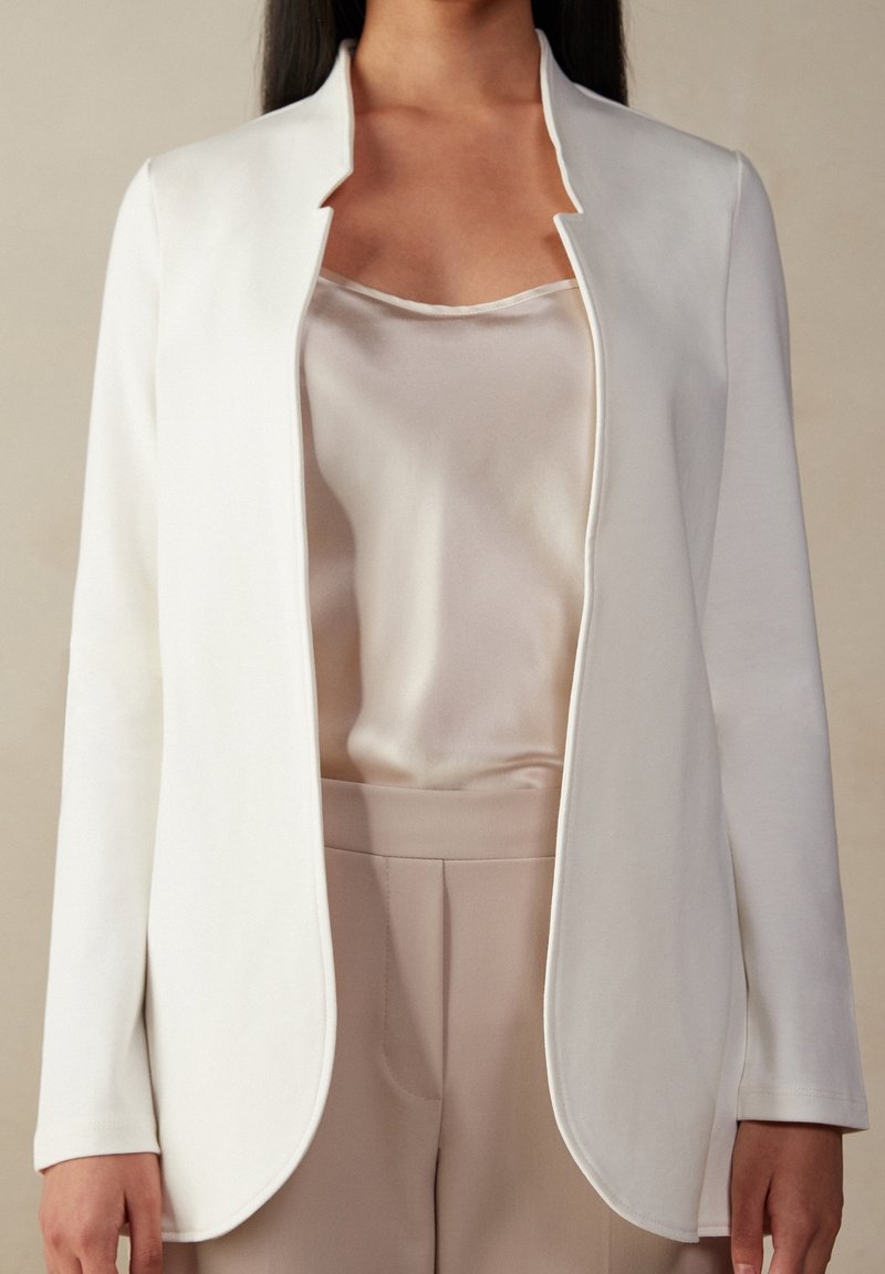 Bílý, lehký blejzr s jemnou texturou, otevřený vpředu a zaobleným lemem, nošený přes lehký saténový top a upravené béžové kalhoty.