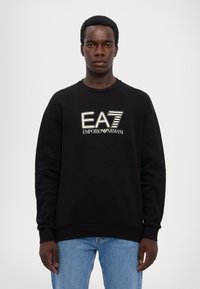 Juodas džemperis su baltu "EA7 Emporio Armani" logo. Dizainas turi gumuotą apykaklę ir rankogalius, o siluetas yra laisvas.