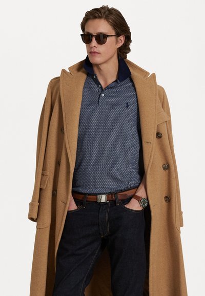 Casaco de lã de camelo, polo azul com padrão, jeans escuros, cinto de couro castanho e óculos de sol; apresenta um design clássico com detalhes de botões.