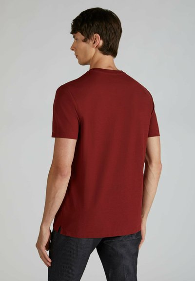 T-shirt in cotone marrone scuro con maniche corte, scollatura rotonda e aperture laterali, mostrato di spalle contro uno sfondo neutro.