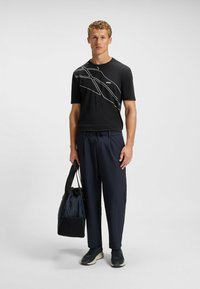 Sort t-shirt med korte ærmer og hvidt abstrakt linjemønster, kombineret med marineblå bukser og en mørk duffel taske. Modellen har mørke sneakers på.