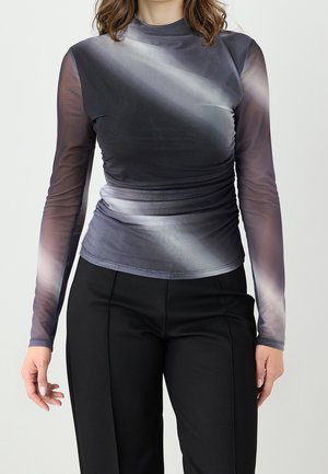 Femme portant un haut ajusté transparent à manches longues avec un motif dégradé foncé et un pantalon noir taille haute sur un fond uni.