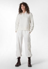 Witte fleece hoodie met een halve ritssluiting, gecombineerd met losse witte cargobroeken met zijzakken en gebundelde enkels. Bessenrode veterschoenen.