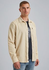 Helle beige Baumwollcordhemd mit durchgehender Knopfleiste, spitzem Kragen und einer Tasche. Über einem schwarzen Grapic-T-Shirt und blauen Jeans getragen.