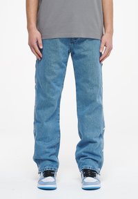Pantalones de mezclilla azul claro con un corte holgado, que cuentan con cierres de botones a presión en los laterales y un diseño de pierna recta, combinados con zapatillas negras y azules.