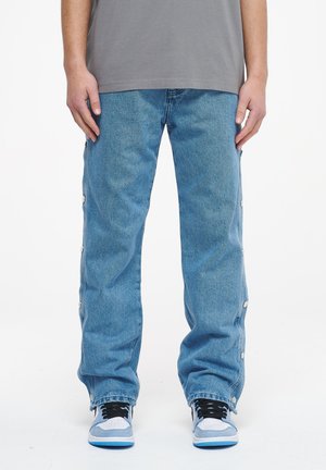 Light blue denim pants with a loose fit, featuring side snap-button closures and a straight leg design, paired with black and blue sneakers.
