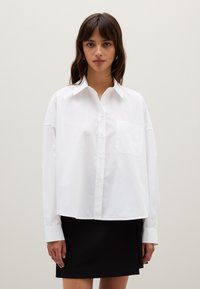 Femme jeune portant une chemise blanche ample à boutons avec une poche poitrine et une jupe noire, devant un fond clair et uni.