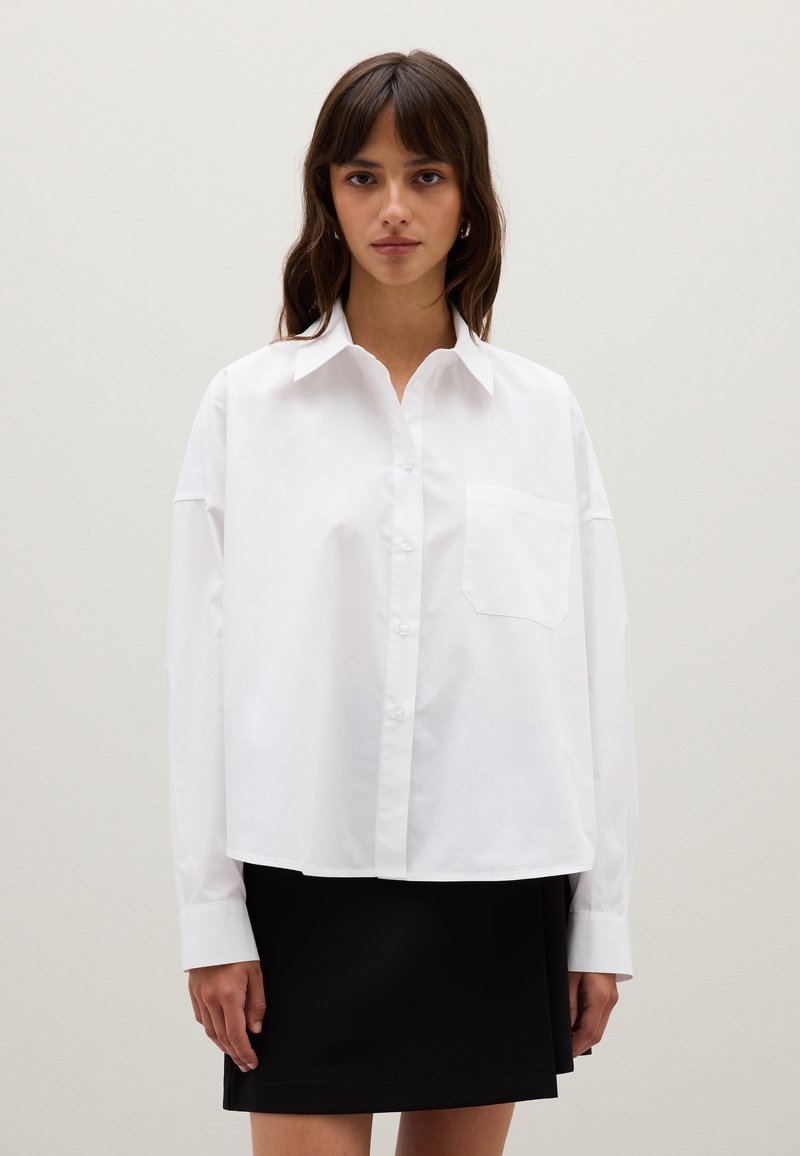 Femme jeune portant une chemise blanche ample à boutons avec une poche poitrine et une jupe noire, devant un fond clair et uni.