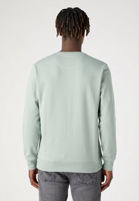 Persona che indossa una felpa verde menta e jeans grigi, in piedi con le spalle rivolte verso l'osservatore su uno sfondo chiaro.