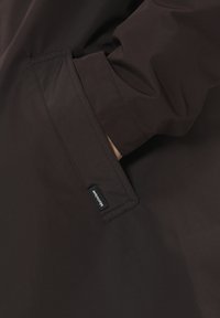 Brązowa kurtka z kieszenią i metką. Materiał wydaje się gładki o matowym wykończeniu, a wzór jest prosty i funkcjonalny.