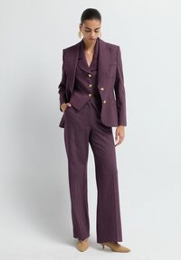 Op maat gemaakt paarse suit met een getailleerde blazer en wijde broekspijen, voorzien van gouden knopen en een gestructureerde stof. Draagt met neutrale hakken.