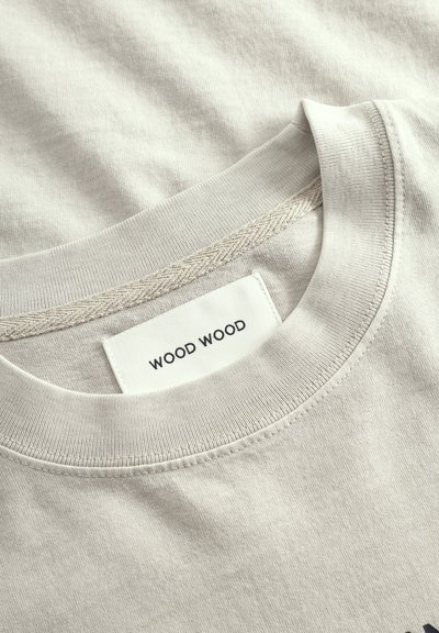 Béžové bavlnené tričko s okrúhlym výstrihom, s textilným štítkom s nápisom "WOOD WOOD" našitým čiernou niťou, hladká textúra a jemné detaily prešívania.