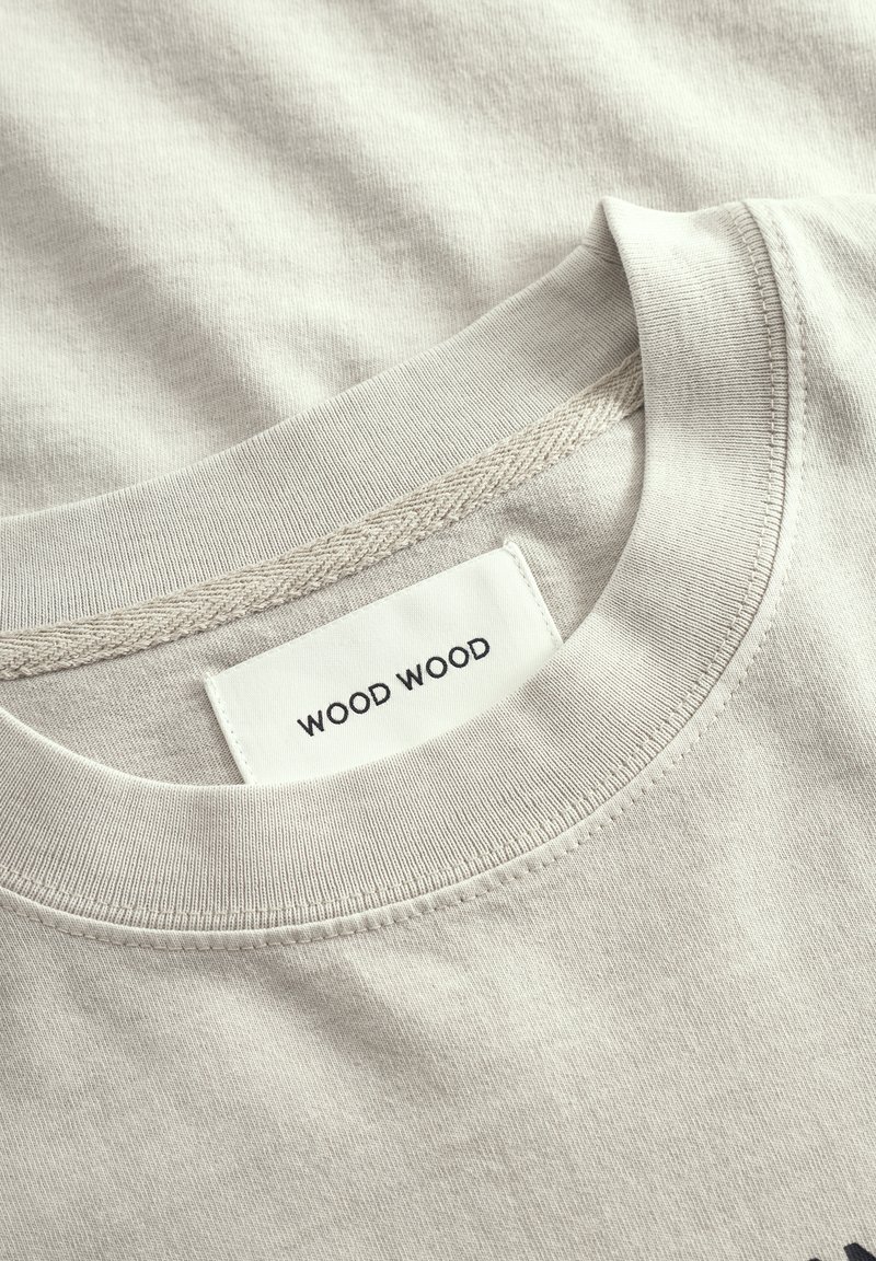 T-shirt en coton beige avec un col rond, présentant une étiquette en tissu portant la mention "WOOD WOOD" brodée en noir, texture lisse et détails de couture subtils.