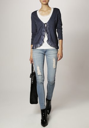 Marineblauwe vest met ruches, gedragen over een wit topje met kanten rand, gecombineerd met blauwe gescheurde jeans en zwarte laarzen met hoge hakken, met een zwarte tas.