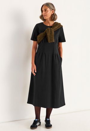 Robe noire midi avec des manches courtes, devant plissé, poches latérales et un châle marron texturé drapé sur l'épaule. Des baskets noires complètent le look.